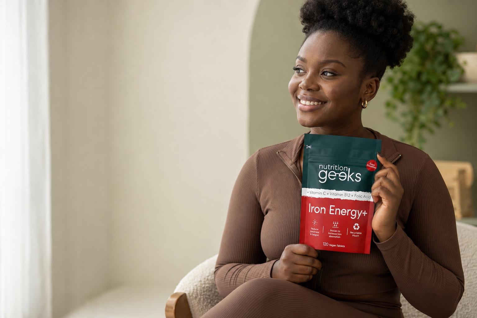 A Ghanaian woman holding a Nutrition Geeks Iron Energy supplement in a warm, sage-toned studio